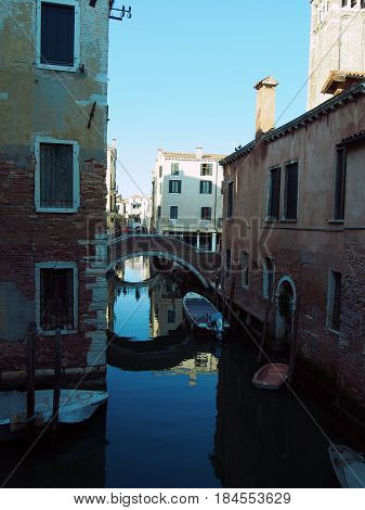 Venice old buidings alongside a small canal whith a bridge reflecting in the water boats and a blue sky