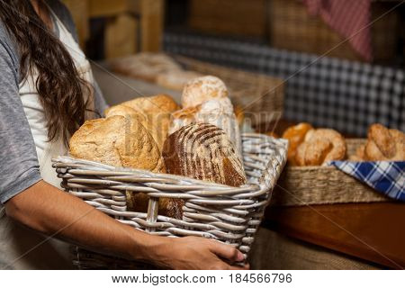 Mid-section of female staff holding wicker basket of various breads at counter in bakery shop