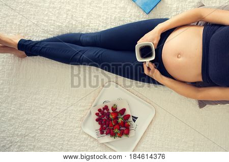 Young Pregnant Woman, Lying In Bed With Smartphone, Book, Coffee And Fruits,