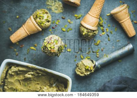 Homemade pistachio ice cream in ceramic mold and metal scooper, with crashed pistachio nuts and waffle cones over dark concrete background, top view, selective focus