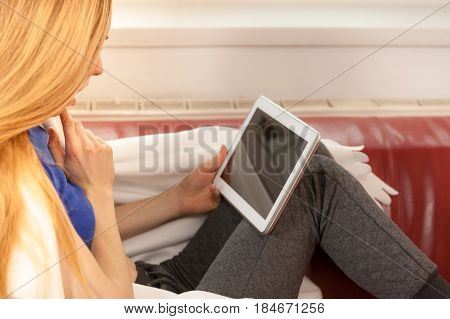 Young woman lying on sofa under warm blanket relaxing using tablet reading news and checking social media having fun during leisure time. Modern relaxation.