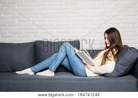 Beautiful Young Woman Reading Book And Lying On Sofa At Home