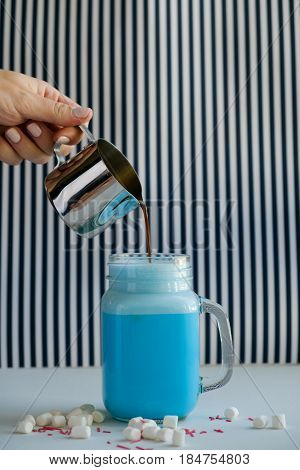 Woman is pouring coffee in cup of colored blue milk on a black-and-white background. Milk shake cocktaill frappuccino. Unicorn coffee unicorn food.