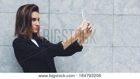 Portrait young businesswomen in black suit using smartphone isolated on background concrete gray wall mockup pretty hipster manager holding mobile gadget girl making selfie photo connect