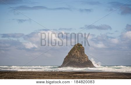Auckland New Zealand - March 2 2017: Wider shot of black sand Karakare Beach under blue cloudy sky with Te Kaka Whakaara Rock (Watchman) in the surf of Tasman Sea.