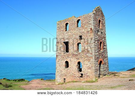 Derelict abandoned Cornish tin mine building on coast path with sea and blue sky in distance
