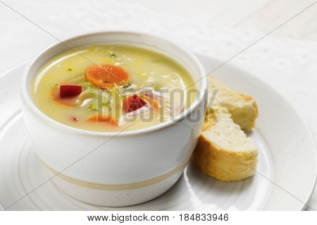soup with healthy vegetables like carrot red pepper and leek in a bright ceramic bowl with bread on a plate white table close up selected focus narrow depth of field