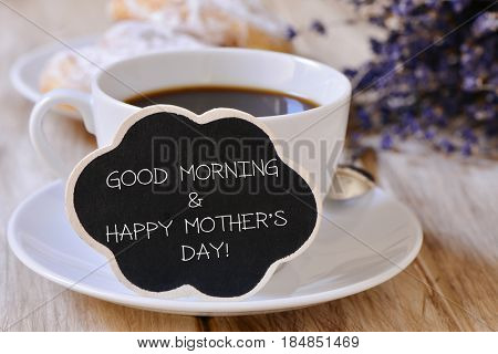 closeup of a black cloud-shaped signboard with the text good morning and happy mothers day written in it, next to a cup of coffee on a table set for breakfast with some viennoiseries in the background