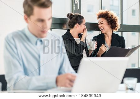 Two young women laughing while talking in the office behind their male colleague