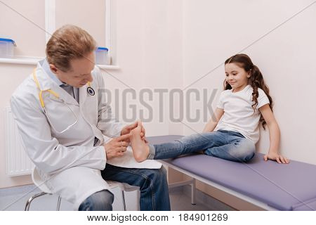 Little toes. Cute nice young girl paying a visit to specialist and sitting on medical bed while the doctor examining her sole