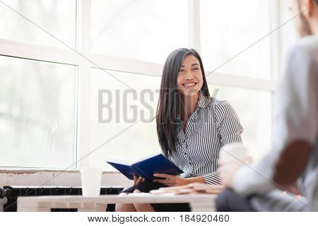 Pretty Asian manager looking at her male colleague with wide smile while having business meeting in office lobby with panoramic windows, portrait shot