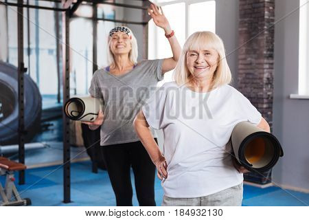Positive emotions. Happy well built senior women standing together and holding yoga mats while enjoying their time in fitness club