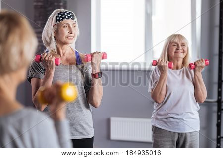 We love fitness. Hard working active elderly women lifting dumbbells and smiling while being in positive mood