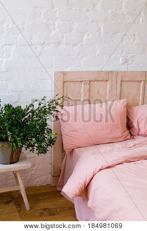 Empty bed with pink pillows and pink cover in the bedroom. White wall wooden floor home flowers with green leaves and small table near the bed. Home interior