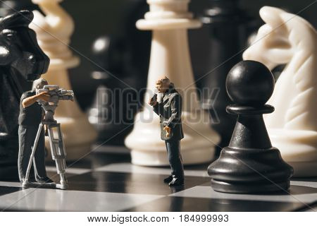 Chess game online broadcast. Filming chess game process. Miniature journalists on chessboard. Chess game process. Chess playing story. Chessmate reporters scene. Black and white figures on game board