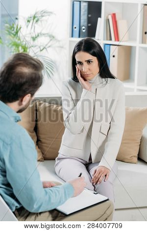 Upset Young Woman Sitting On Couch And Looking At Psychotherapist With Clipboard