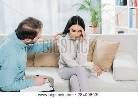 Psychotherapist Writing On Clipboard And Looking At Young Upset Woman Sitting On Couch