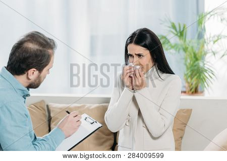 Upset Young Woman Crying And Looking At Bearded Psychotherapist With Clipboard