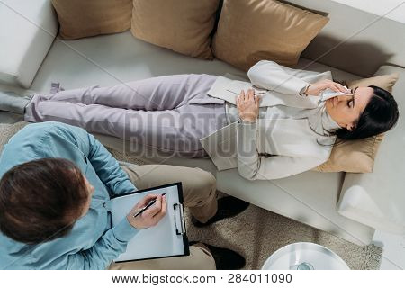Overhead View Of Psychotherapist Writing On Clipboard And Young Patient Crying On Couch