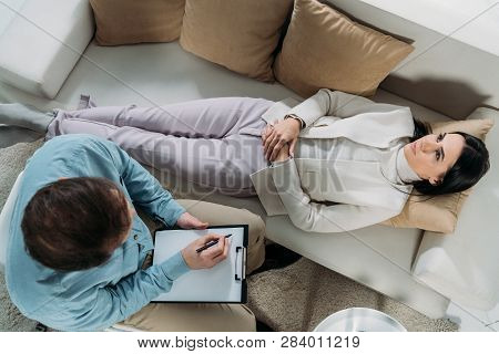 Overhead View Of Psychotherapist Writing On Clipboard And Upset Young Patient Lying On Couch