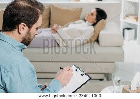 Bearded Psychotherapist Writing On Clipboard While Upset Patient Lying On Couch