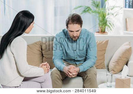 Young Psychotherapist Talking With Upset Bearded Man Sitting On Couch