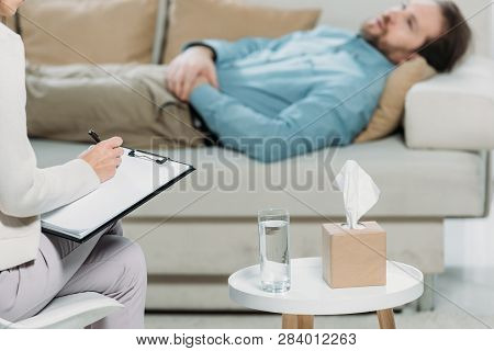 Cropped Shot Of Psychotherapist Writing On Clipboard While Bearded Man Lying In Couch