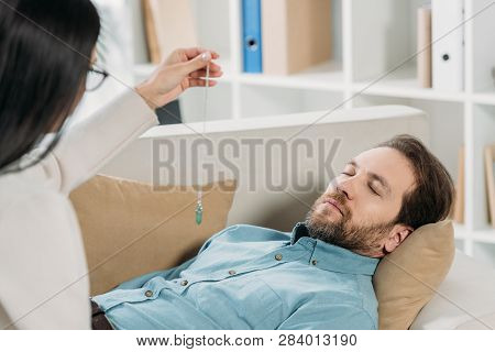 Young Woman With Pendulum Hypnotising Bearded Man With Closed Eyes Lying On Couch