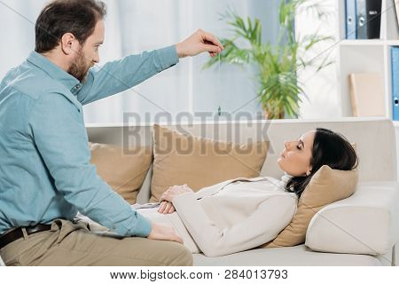 Side View Of Bearded Hypnotist Holding Pendulum And Hypnotising Young Woman With Closed Eyes Lying O