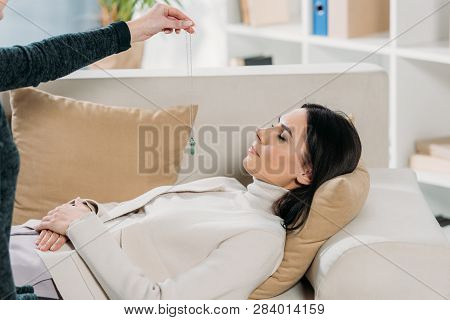 Cropped Shot Of Hypnotist With Pendulum Hypnotizing Young Woman Lying On Couch