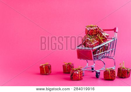 Small Shopping Cart With Presents On Pink Background. Trolley From A Supermarket Full Of Red Gift Bo