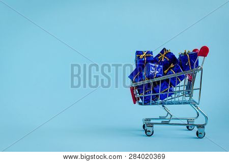 Small Shopping Cart With Presents On Blue Background. Trolley From A Supermarket Full Of Blue Gift B