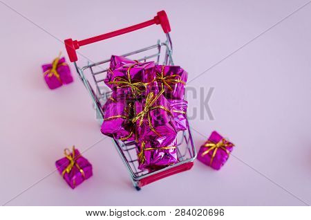 Small Shopping Cart With Presents On Pink Background. Trolley From A Supermarket Full Of Purple Gift