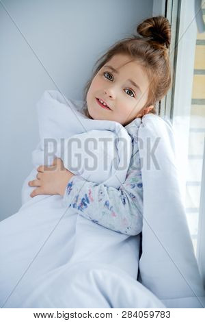 A little girl sittiing on a windowsill on a pillow covered with a blanket. Childhood. Kid's fashion.
