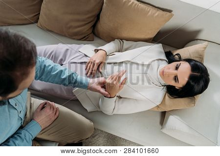 Overhead View Of Hypnotist Holding Wrist Of Young Woman Lying In Couch