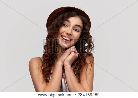 Beautiful Young Joyful Woman Keeps Hands Together, Has Brown Eyes Full Of Happiness, Stands Against 