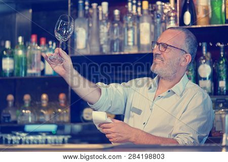 Elderly Bartender Cleans The Glass. A Handsome Bartender Polishes A Glass Of Wine Glasses. The Conce