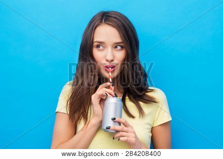 people concept - smiling young woman or teenage girl in yellow t-shirt drinking soda from can through paper straw over bright blue background