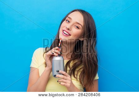people concept - smiling young woman or teenage girl in yellow t-shirt drinking soda from can through paper straw over bright blue background