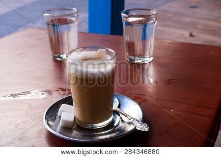 Traditional Moroccan Coffee With Milk In A Simple Glass. Plate, Ornate Spoon And Sugar Cubes. Dirtly