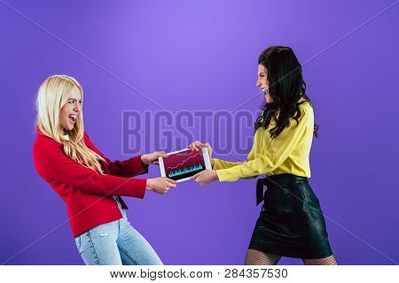 Angry Young Women Fighting For Digital Tablet With Traiding App On Screen On Purple Background