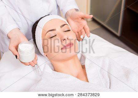 Beautician Holding Cosmetic Cream Bottle And Puting It On Woman Face At Beauty Salon