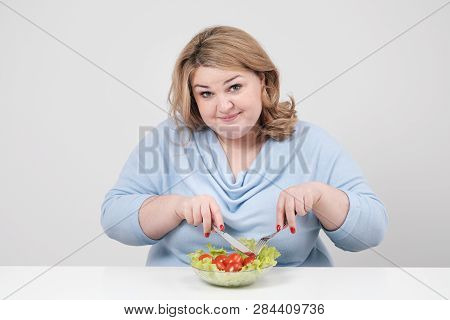 Young Lush Fat Woman In Casual Blue Clothes On A White Background At The Table And Eats A Vegetable 