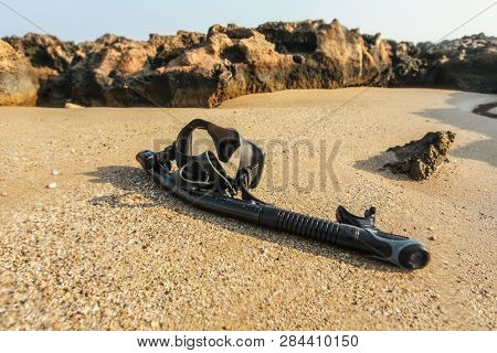 Black Snorkel And Diving Mass On Beach Sand, Small Rocks In Background. Shot Through Wet Lens (drops