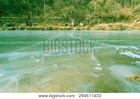 Low Angle View Of Cracks In Ice On Surface Of River With Treed Shoreline And Country Road In Backgro