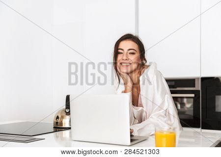 Photo of smiling woman 20s wearing housecoat using laptop in kitchen