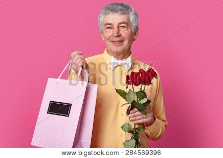 Handsome Wrinkled Mature Grey Hiared Man Ready For Party, Wants To Give Gift Bag And Red Flowers To 