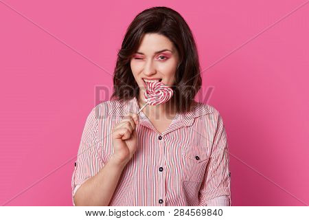 Photo Of Attractive Young Woman With Dark Haired Woman With Bright Makeup, Bites Candy, Blinks Eye, 