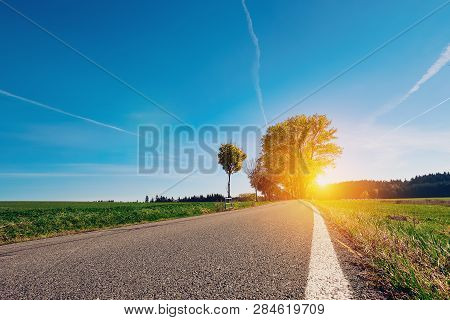 Long Empty Asphalt Country Road With White Side Line And Green Grass Stratching Off To A Lone Tree A