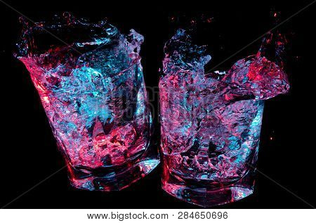 Two Rocks Glasses Clinking In Toast While Glowing Colorful Liquid Splashes Out On A Black Background